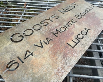 May include: A weathered metal sign with the text "GOOSYS NEST S14 VIA MONTE BONELLI LUCCA" cut out. The sign has a rustic, aged appearance with a mix of brown and gray tones, suggesting outdoor use.
