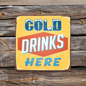Könnte beinhalten: Ein Vintage-Schild aus Metall mit gelbem Hintergrund, rotem Sechseck und blauem Text. Das Schild lautet "COLD DRINKS HERE" mit schneeähnlichen Akzenten auf dem Wort "COLD".
