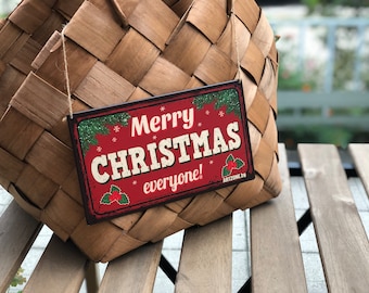 May include: A rectangular Christmas sign with a red background and the words "Merry CHRISTMAS everyone!" in white. The sign has a distressed, vintage look with green accents and holly illustrations. It hangs from a woven basket.