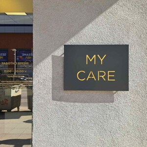 Pode incluir: Uma placa preta com letras douradas que lê "MY CARE" está montada em uma parede branca. A placa está na frente de uma porta de vidro com um reflexo de um prédio e uma placa com texto em um idioma estrangeiro.