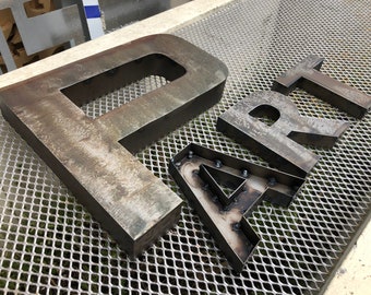 May include: Metal letters spelling "ART" in a rustic, industrial style. The letters are thick and have a weathered, metallic finish. The letter "A" has visible bolts and welds, while the others are solid.
