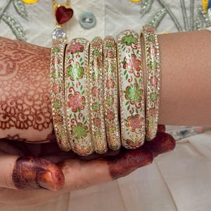 Puede incluir: Un conjunto de brazaletes blancos con detalles dorados y diseños florales en rosa y verde. Los brazaletes están decorados con pequeños diamantes de imitación transparentes.