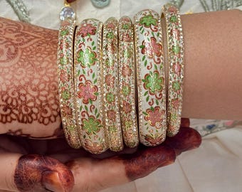 Könnte beinhalten: Ein Set weißer Armreifen mit goldenen Akzenten und floralen Designs in Rosa und Grün. Die Armreifen sind mit kleinen, klaren Strasssteinen verziert.