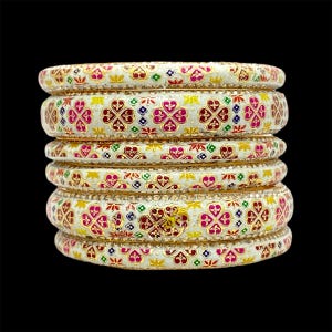 May include: Stack of six white bangles with intricate floral patterns in red, pink, gold, green, and blue. Each bangle has a gold-colored border with small, sparkling embellishments. The bangles are stacked against a black background.