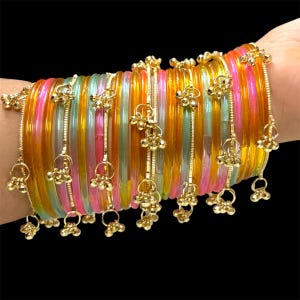 May include: A stack of colorful bangles in shades of orange, pink, green, and clear. Each bangle is adorned with dangling gold-colored charms. The bangles are displayed against a black background.