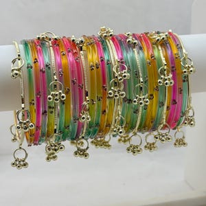 以下が含まれることがあります： ピンク、緑、黄色、オレンジの色合いのカラフルなバングルのコレクション。各バングルには、小さな金色のチャームがぶら下がっています。バングルは白い表面に展示されており、鮮やかな色と繊細なディテールが際立っています。