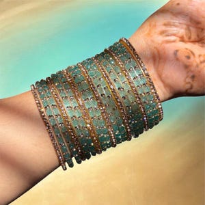 May include: A collection of teal and gold bangles adorns a wrist. The bangles feature intricate floral patterns and are embellished with small, sparkling accents. The wrist is set against a gradient background of teal and gold.