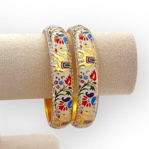 Puede incluir: Dos brazaletes dorados con intrincados diseños florales y animales en rojo, azul y verde sobre un fondo blanco. Los brazaletes se exhiben en un soporte beige, mostrando su detallada artesanía y colores vibrantes.