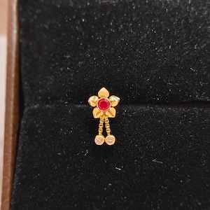 Op de afbeelding: Een goudkleurige neusstud met een bloemdesign. De bloem heeft een centrale rode edelsteen en twee bungelende heldere edelstenen. De sieraden zijn geplaatst tegen een donkere achtergrond. De bloem heeft vijf bloemblaadjes.