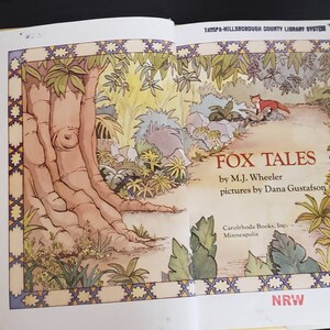 May include: A book titled "Fox Tales" by M.J. Wheeler with illustrations by Dana Gustafson. The cover features a fox walking on a path through a lush green forest.