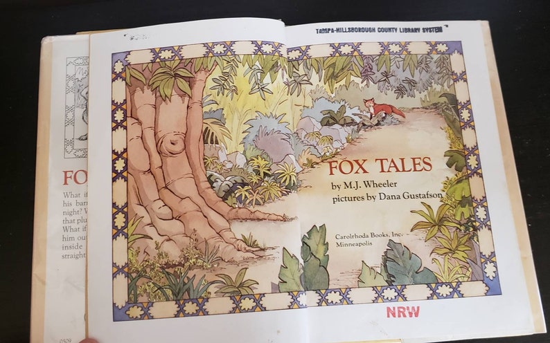 May include: A book titled "Fox Tales" by M.J. Wheeler with illustrations by Dana Gustafson. The cover features a fox walking on a path through a lush green forest.