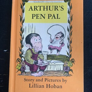 May include: An orange book cover with a title that reads "Arthur's Pen Pal". The cover features illustrations of two monkeys, one sitting and writing in a notebook and the other jumping rope. The text "Story and Pictures by Lillian Hoban" is printed below the illustrations.