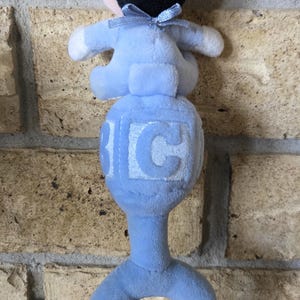 May include: A blue plush baby rattle with a Mickey Mouse-like figure on top. The rattle has a ring handle and a cube with the letter "C" on it. The figure has black ears and a blue bow.