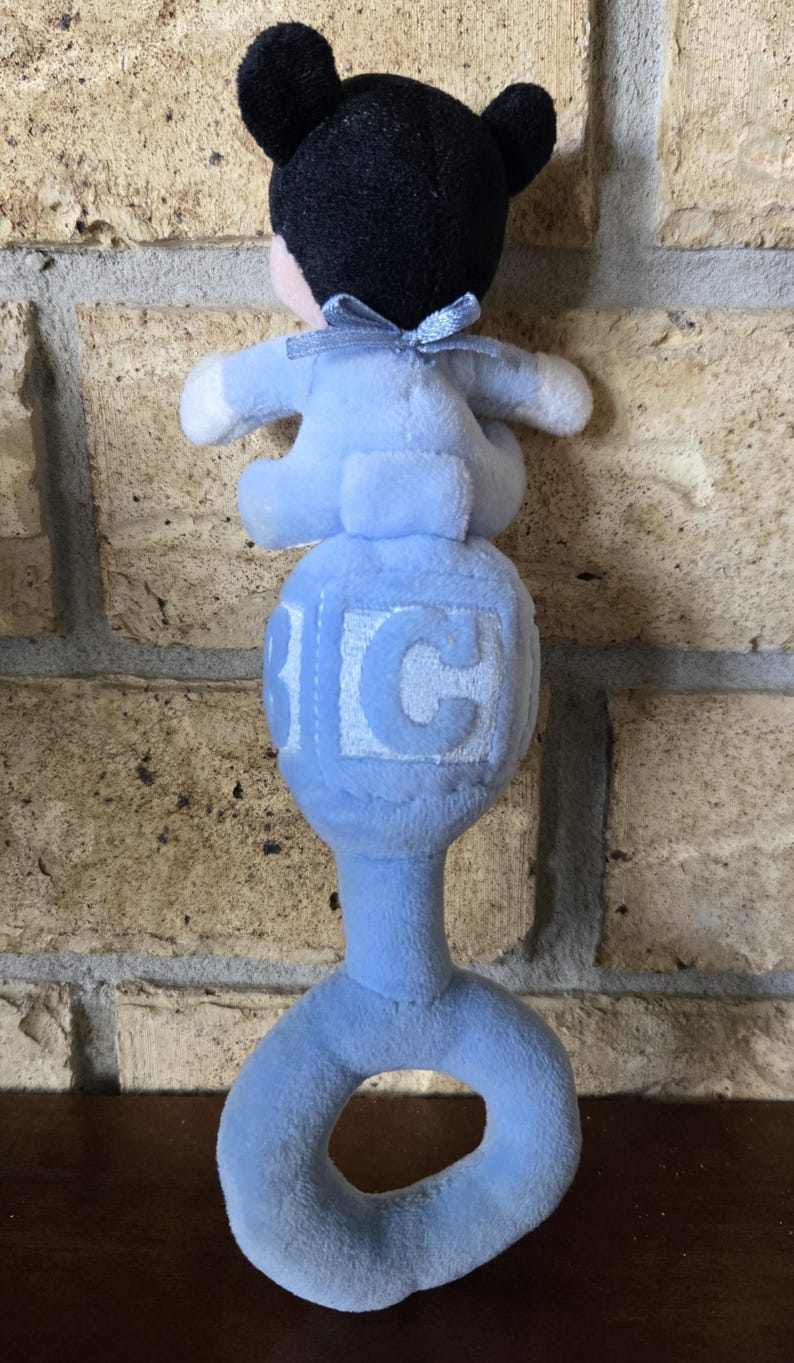 May include: A blue plush baby rattle with a Mickey Mouse-like figure on top. The rattle has a ring handle and a cube with the letter "C" on it. The figure has black ears and a blue bow.