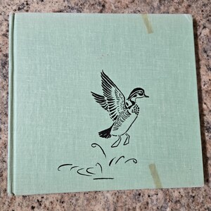 May include: A light green book with a black illustration of a duck in flight. The duck has detailed wing feathers and is positioned above a stylized depiction of grass. The book has a textured cover and is square in shape.