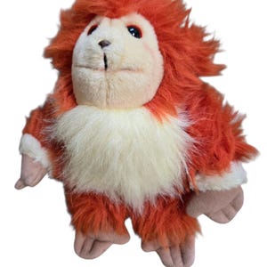 May include: A plush toy in the shape of a furry creature. The toy has orange fur around its head and body, with a cream-colored chest. It has a light brown face, hands, and feet. The toy has a friendly expression.