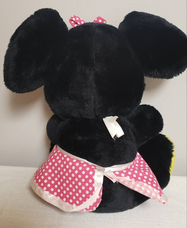 May include: A black plush toy with large ears and a pink polka dot bow. The toy is wearing a pink and white polka dot skirt. The toy is sitting down.