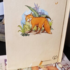 May include: The back cover of a children's book featuring a cartoon fox with orange fur standing in a green forest. The book title is "The Fox and the Tale".