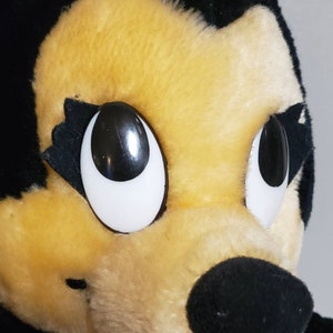 May include: Close-up of a plush toy character with a black head, ears, and nose, and a yellow face. The character has large, white-rimmed eyes and a pink bow with white polka dots. The character is wearing a pink and white polka dot dress.