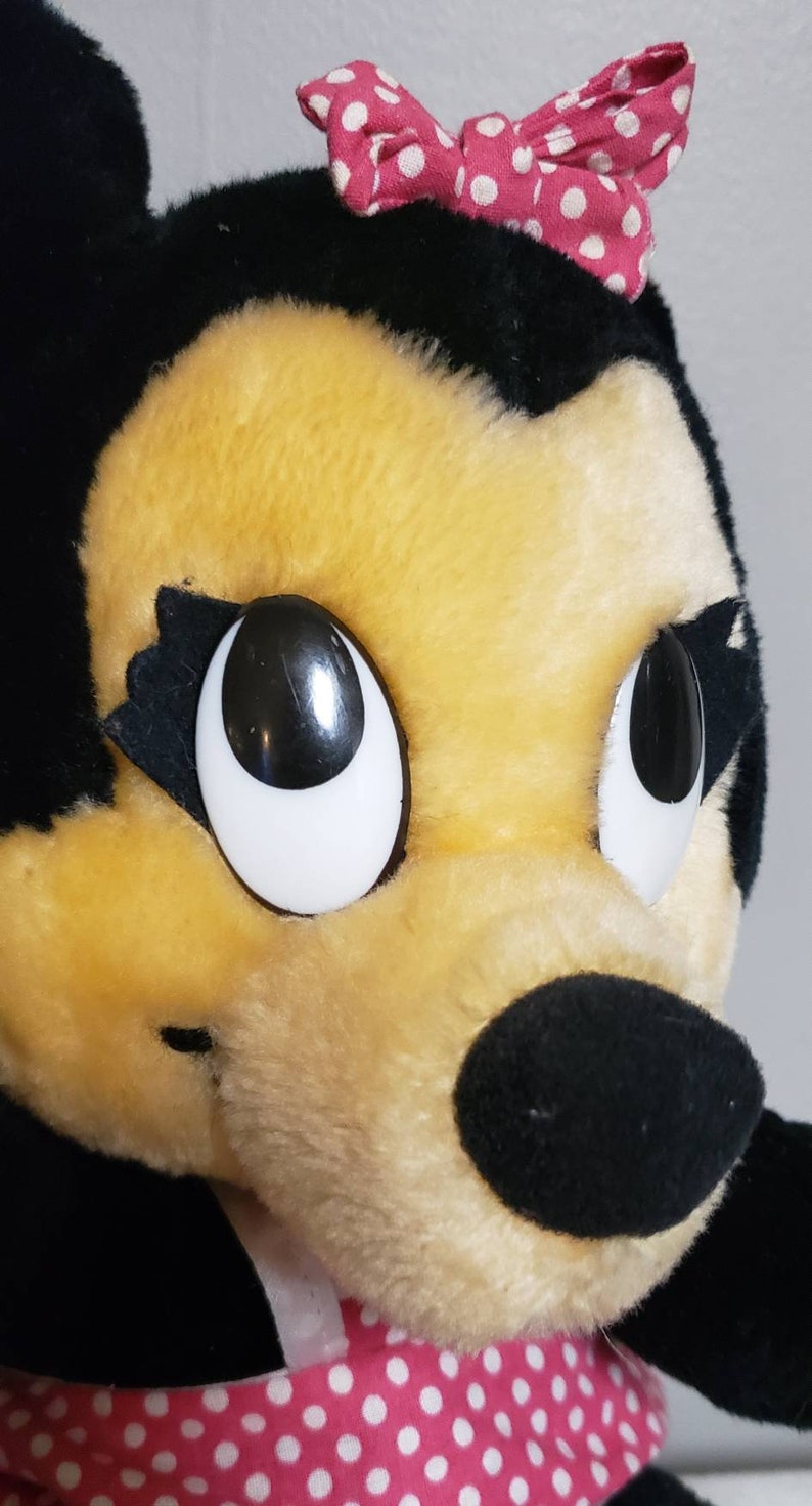 May include: Close-up of a plush toy character with a black head, ears, and nose, and a yellow face. The character has large, white-rimmed eyes and a pink bow with white polka dots. The character is wearing a pink and white polka dot dress.