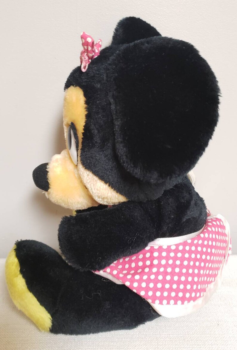 May include: A plush Minnie Mouse doll, featuring black fur, a yellow muzzle, and a pink polka-dot skirt. A pink bow adorns the top of the head. The doll is in a seated position, showcasing its classic design.