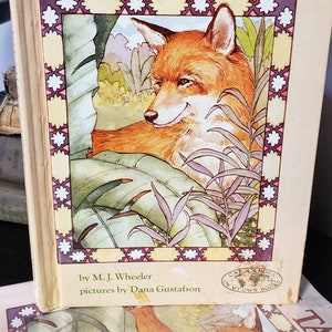 May include: A vintage children's book titled "Fox Tales" by M.J. Wheeler with illustrations by Dana Gustafson. The cover features a red fox sitting in a lush green jungle setting.
