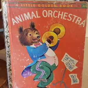 May include: A Little Golden Book titled "Animal Orchestra" with a red cover featuring a cartoon bear wearing a blue suit and playing cymbals. The bear is smiling and has a red bow tie. The book has a gold spine with a decorative pattern.