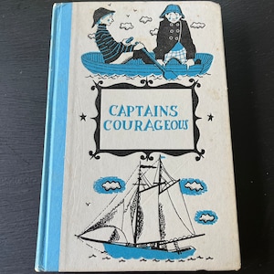 May include: A vintage hardcover book with a blue and white cover featuring a black and white illustration of a sailboat and two children in a rowboat. The title of the book is "CAPTAINS COURAGEOUS".