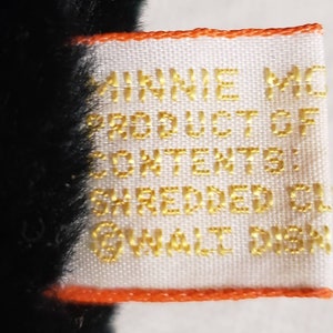May include: Close-up of a white tag with gold text and an orange border. The tag reads "MINNIE MO... PRODUCT OF... CONTENTS: SHREDDED CL... &copy;WALT DISN..." The tag is attached to a black plush toy.