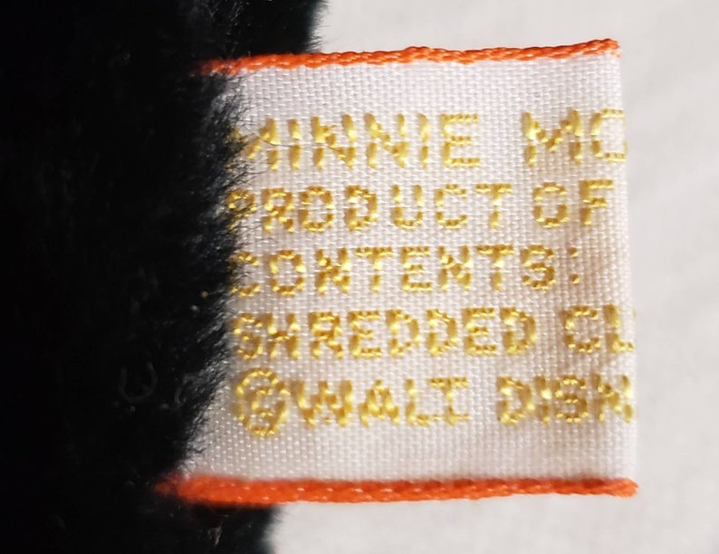 May include: Close-up of a white tag with gold text and an orange border. The tag reads "MINNIE MO... PRODUCT OF... CONTENTS: SHREDDED CL... &copy;WALT DISN..." The tag is attached to a black plush toy.