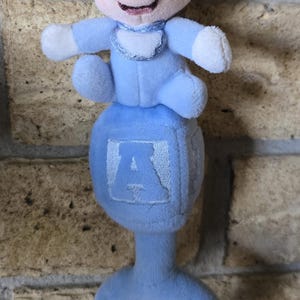 May include: A plush baby rattle featuring a Mickey Mouse figure in a light blue outfit. The rattle has a blue handle and a cube with the letter "A". The Mickey Mouse figure has black ears, a pink face, and white gloves.