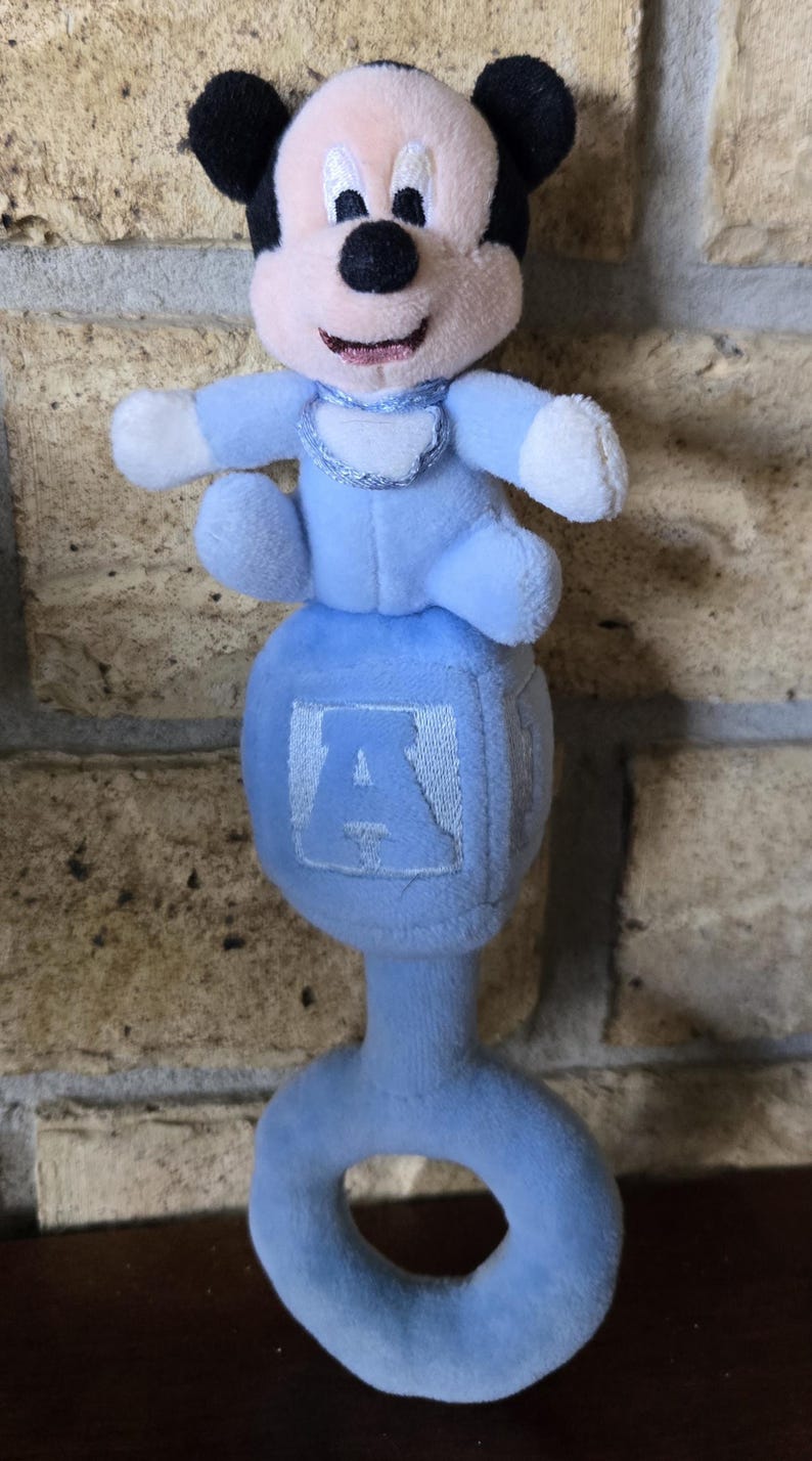 May include: A plush baby rattle featuring a Mickey Mouse figure in a light blue outfit. The rattle has a blue handle and a cube with the letter "A". The Mickey Mouse figure has black ears, a pink face, and white gloves.