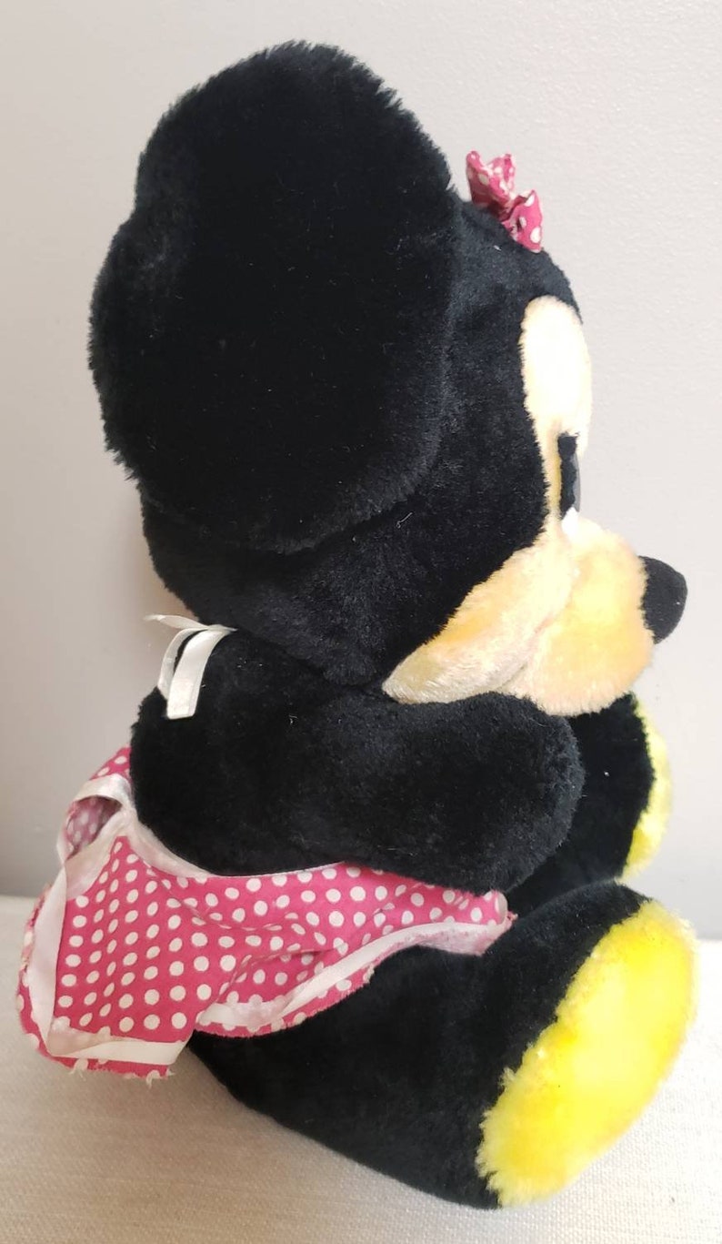 May include: A plush Minnie Mouse doll, primarily black with yellow accents on the feet and face. She wears a pink polka-dot dress and a matching bow. The doll is soft and cuddly, perfect for children.