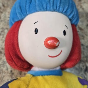 May include: A plush clown doll with a white face, red hair, and a blue hat. The clown has a red nose and a smiling mouth. The doll is wearing a yellow shirt with blue and purple polka dots. A classic toy.
