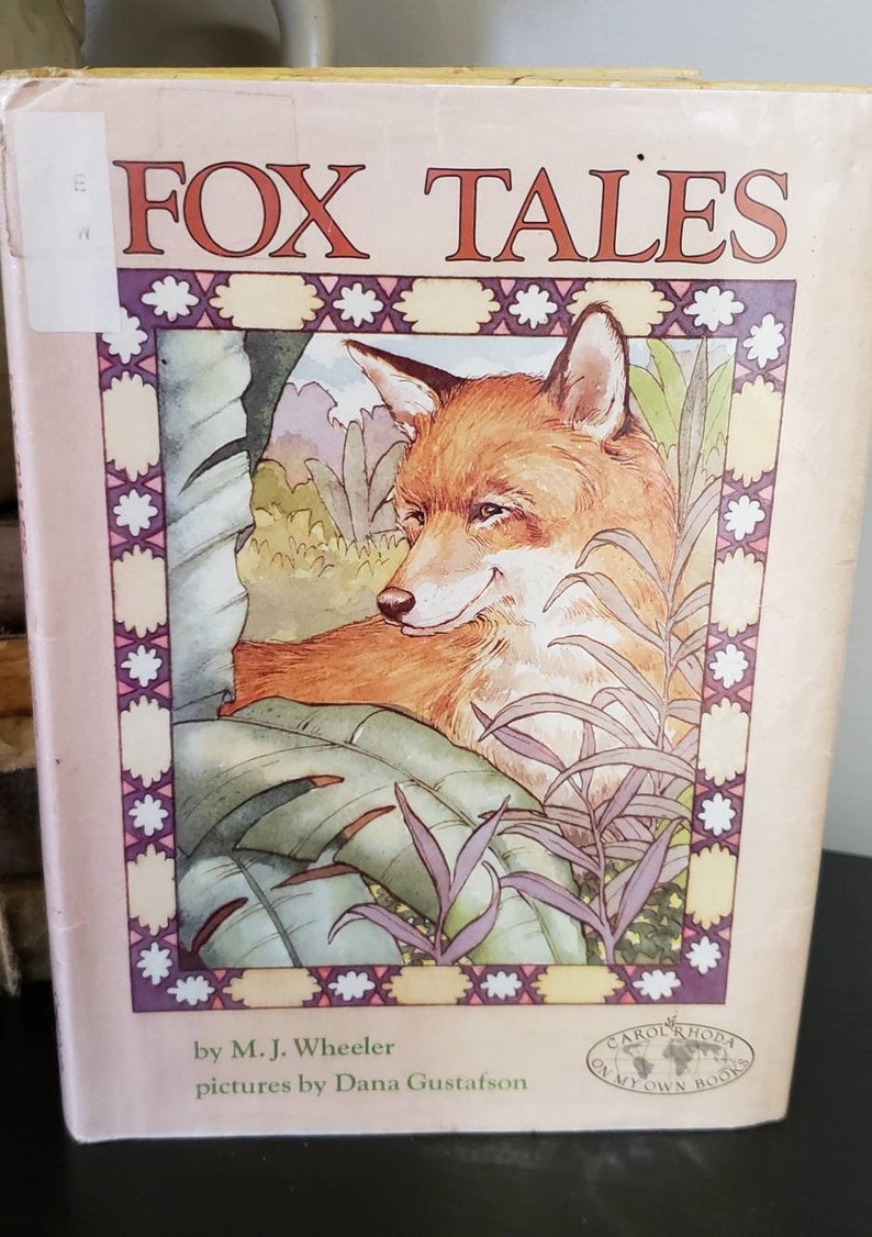May include: A children's book titled "Fox Tales" by M.J. Wheeler with illustrations by Dana Gustafson. The cover features a red fox peeking out from behind green leaves.