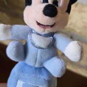 May include: A plush Mickey Mouse toy in a light blue outfit. The toy has black ears, a pink nose and mouth, and a white bib. The toy is sitting on a blue block with the letter "A" embroidered on it.