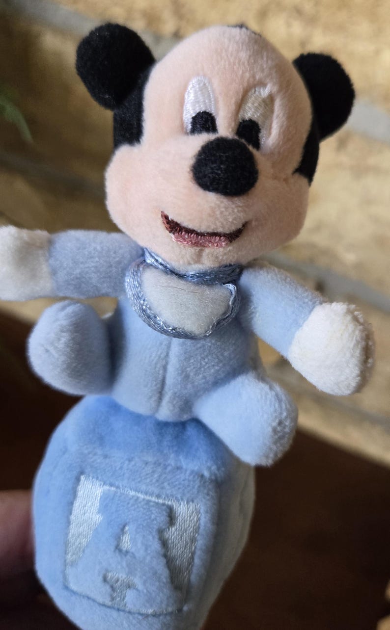 May include: A plush Mickey Mouse toy in a light blue outfit. The toy has black ears, a pink nose and mouth, and a white bib. The toy is sitting on a blue block with the letter "A" embroidered on it.