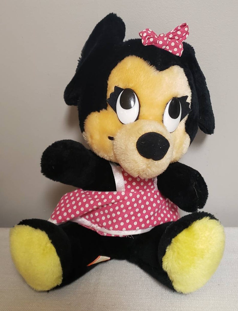 May include: A plush dog toy with black fur, a yellow face, and large, expressive eyes. It wears a pink polka-dot dress and a matching bow. The toy has yellow paws and is sitting upright.