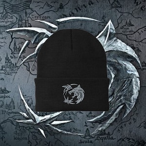 May include: Black beanie with a gray embroidered wolf's head logo on the front. The logo is a stylized wolf's head with a crescent moon and a stylized tree behind it. The beanie is on a background of a gray map with a silver dragon design.