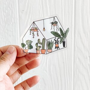 May include: A clear sticker of a greenhouse with a variety of plants, including cacti, succulents, and leafy plants. The greenhouse is decorated with hearts and a light bulb.
