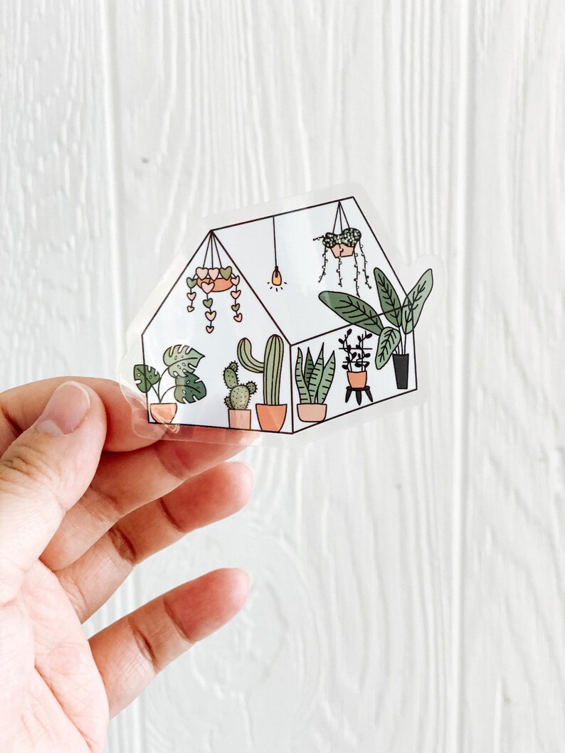 May include: A clear sticker of a greenhouse with a variety of plants, including cacti, succulents, and leafy plants. The greenhouse is decorated with hearts and a light bulb.