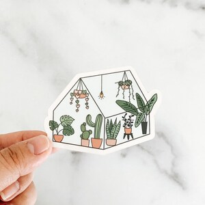 May include: A white sticker with a black outline of a greenhouse filled with various plants, including cacti, succulents, and leafy plants. The greenhouse has a light bulb and a hanging plant with pink hearts.