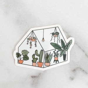 May include: A sticker of a greenhouse with a variety of plants, including cacti, succulents, and leafy plants. The greenhouse is decorated with pink hearts and a light bulb.