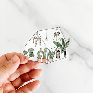 May include: A clear sticker with a line drawing of a greenhouse filled with various plants, including a cactus, a snake plant, and a monstera plant. The greenhouse has a light bulb and hanging plants with hearts.