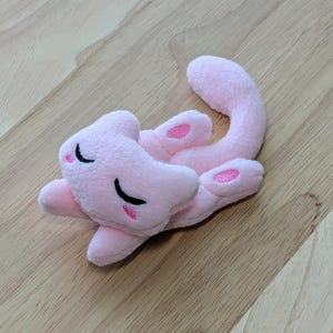 Puede incluir: Juguete de peluche rosa con forma de gato con los ojos cerrados y una nariz rosa. El gato está acostado de espaldas con la cola enroscada alrededor de su cuerpo.