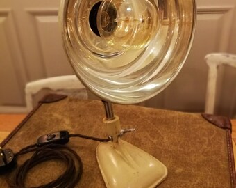 Könnte beinhalten: Eine weiße Vintage-Tischlampe mit einem großen, klaren Glasschirm und einer runden Glühbirne. Die Lampe hat einen flexiblen Arm und einen Sockel mit einem Kabel und einem Stecker.