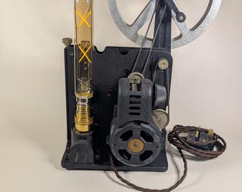 Könnte beinhalten: Eine umgebaute Vintage-Filmprojektor-Lampe mit schwarzem Metallsockel, einer großen silberfarbenen runden Spule und einer hohen, bernsteinfarbenen röhrenförmigen Edison-Glühbirne mit gelbem Kreuz-Glühfaden, verbunden durch ein braunes Textilkabel.