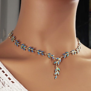 Pode incluir: Um colar dourado com um design de folhas e detalhes em opala azul. O colar é usado ao redor do pescoço e tem uma aparência delicada e elegante. As folhas são dispostas em um padrão simétrico.