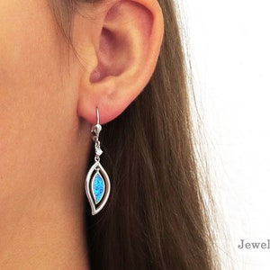 May include: Silver-toned earrings with a leaf-shaped design. Each earring features a vibrant blue opal stone set within a delicate, openwork frame. The earrings are shown on a person's ear, with a lever-back closure. The website JewelryByPoulopoulos.com is visible.