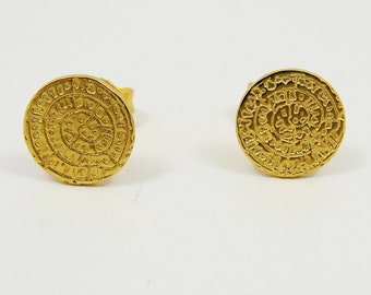 May include: A pair of gold-toned earrings with a circular design featuring intricate patterns and symbols. The earrings are made of metal and have a post-back closure.